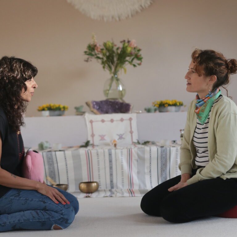 Two women facing each other in a meditative pose on cushions, with a table containing flowers and spiritual objects in the background.