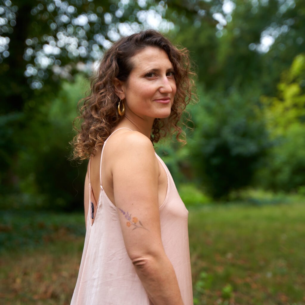 Woman with curly hair wearing a pink dress standing in a garden setting, looking over her shoulder.